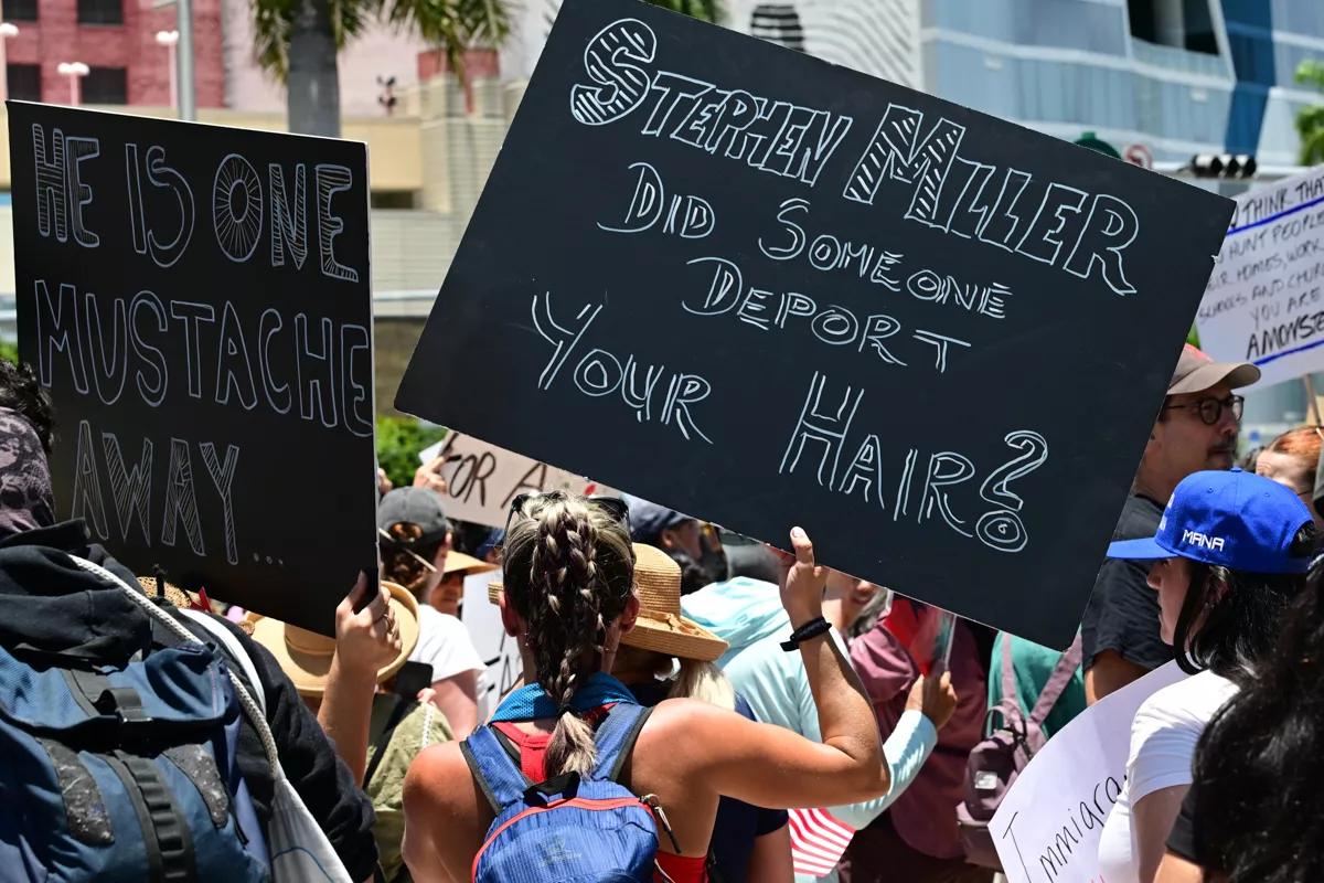 A protester with her back towards the camera holds a black sign that reads, "Stephen Miller did someone deport your hair?"