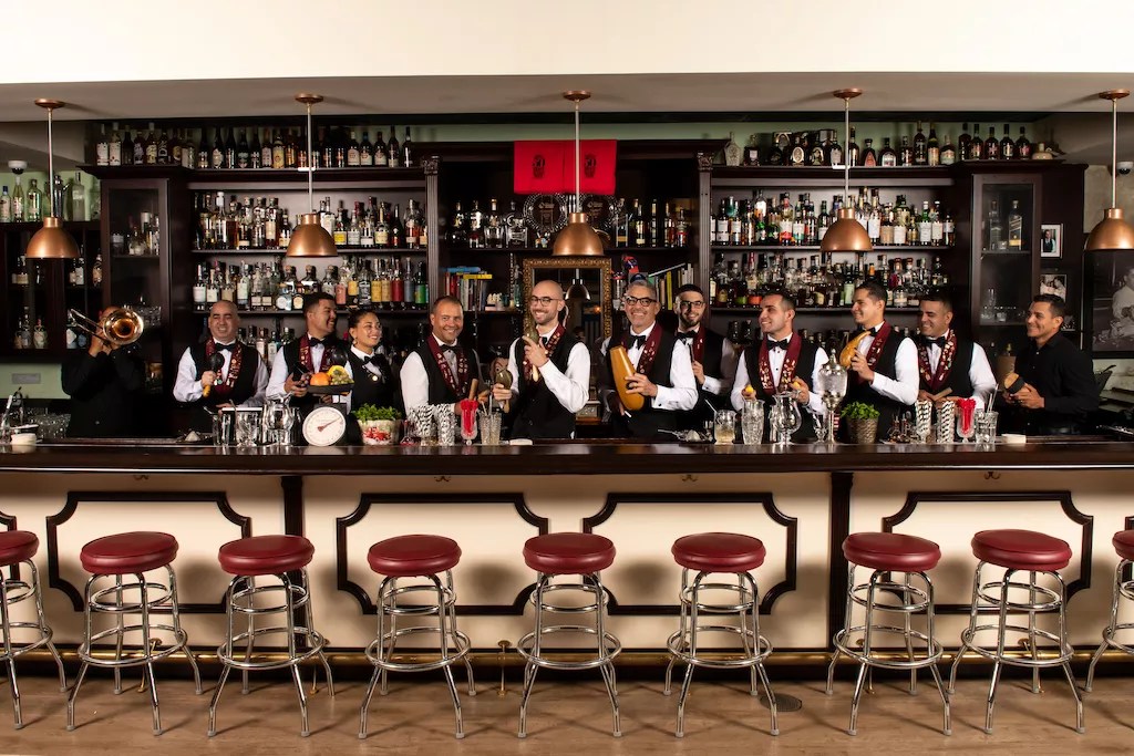 Bartenders in white shirts and black vests smile from behind at a bar.