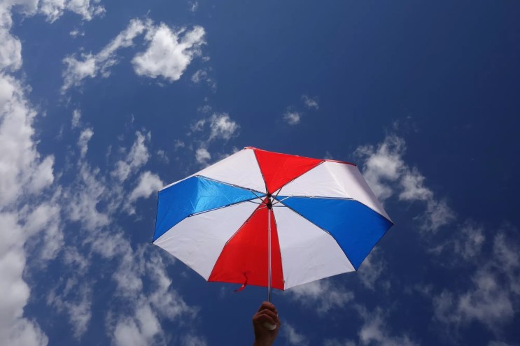 A red, white, and blue umbrella held up against a mostly clear sky with scattered clouds