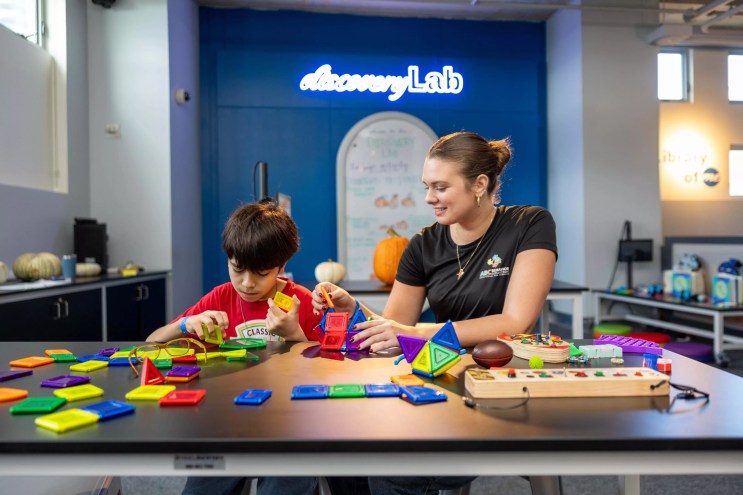 A child and a woman play with puzzles and toys on a table. A neon sign behind them reads, "discoverLab."