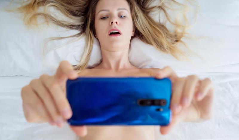 Young sexy woman posing in front of smartphone in her hand and working as webcam model