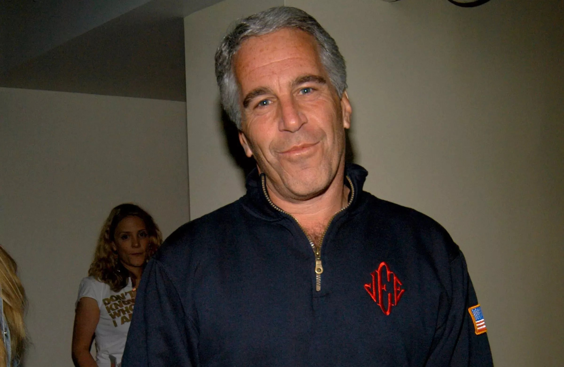 Jeffrey Epstein looking at the camera wearing the blue quarter zip sweatshirt with red monogrammed initials